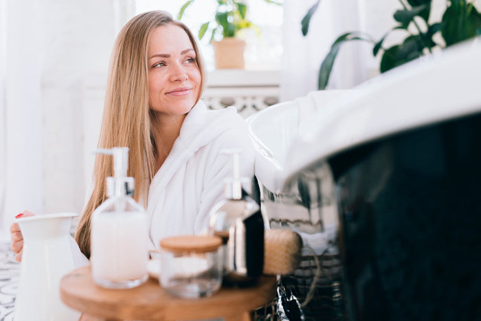 Woman in a white robe enjoying a self-care routine in the bathroom with skincare and beauty products by the bathtub.