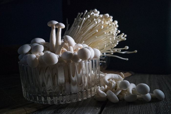 Close-up of fresh white mushrooms in a glass bowl with enoki mushrooms in the background on a wooden table.