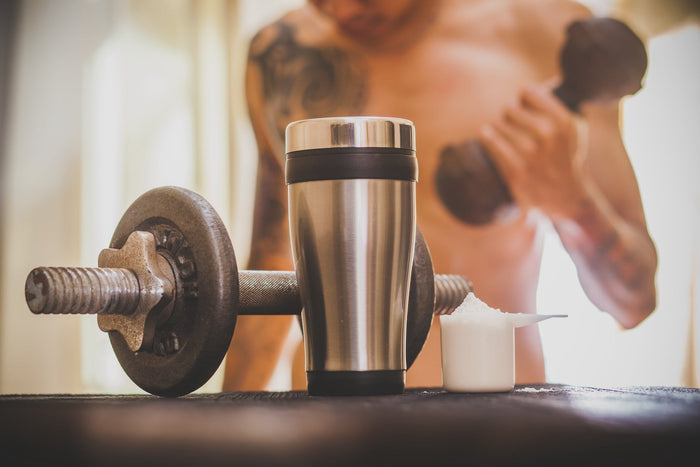 Pre workout coffee and supplements alongside gym equipment with a fit man lifting  dumbbells in the background.