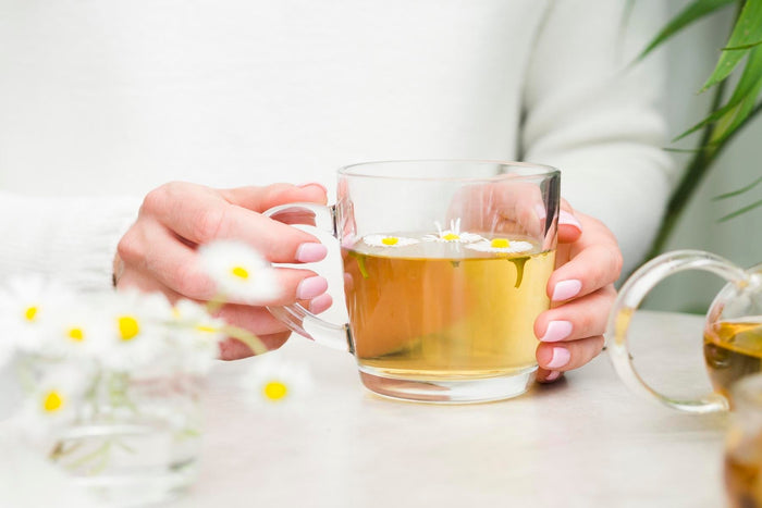 Woman holding a glass cup of herbal tea with chamomile flowers on top, healthy natural tea drink on table.