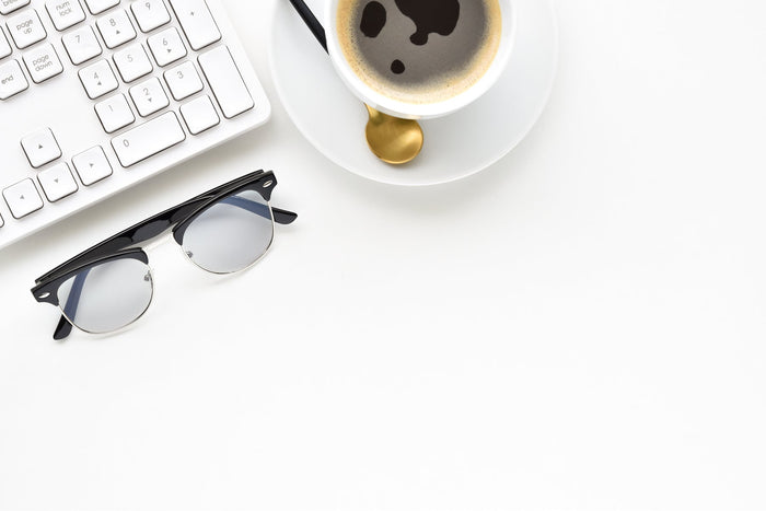 A cup of black coffee, stylish sunglasses, and a white computer keyboard on a clean desk.