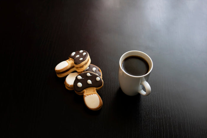  Mushroom coffee in a white cup with mushroom shaped cookies on a dark wooden table. 