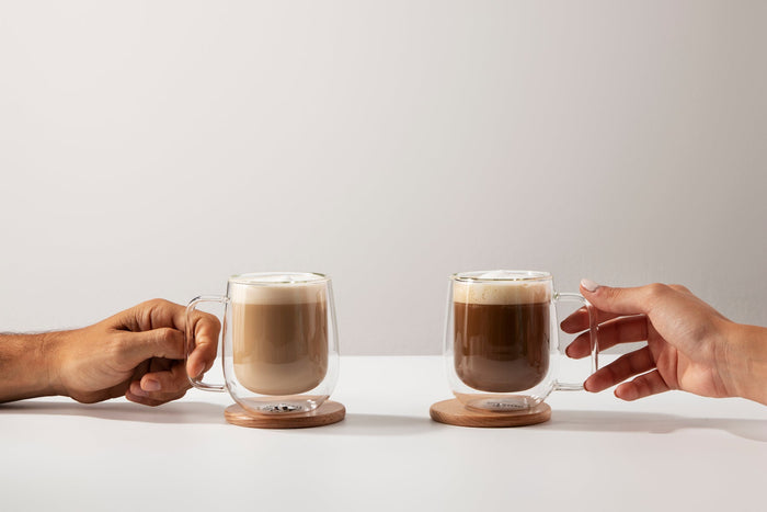Two hands holding glass mugs of coffee with frothy tops, placed on wooden coasters against a light background.